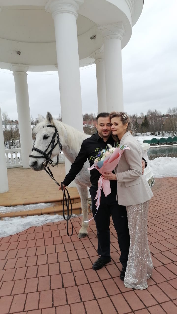 На фотографии пара, мужчина держит белую лошадь за повод, другой рукой обнимает свою девушку, невесту